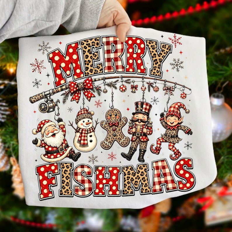 May include: A white t-shirt featuring a graphic with the text MERRY FISHMAS. The design includes a fishing rod hanging ornaments like a snowman, gingerbread man, nutcracker, and elf, with Santa Claus below, all in red, white, and leopard print.