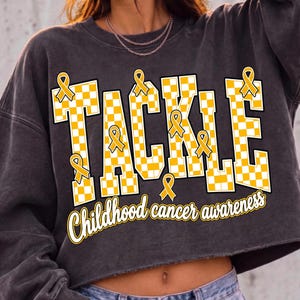 May include: A charcoal gray cropped sweatshirt featuring the word TACKLE in large, yellow and white checkered block letters. Several small yellow awareness ribbons are placed around the letters, with the text Childhood cancer awareness below.