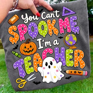 May include: A dark gray t-shirt featuring a colorful graphic with the text "You Can't SPOOK ME I'm a TEACHER". The design includes a jack-o'-lantern, a globe, a ghost with flower patterns, scissors, a pencil, an eraser, and a triangle ruler.