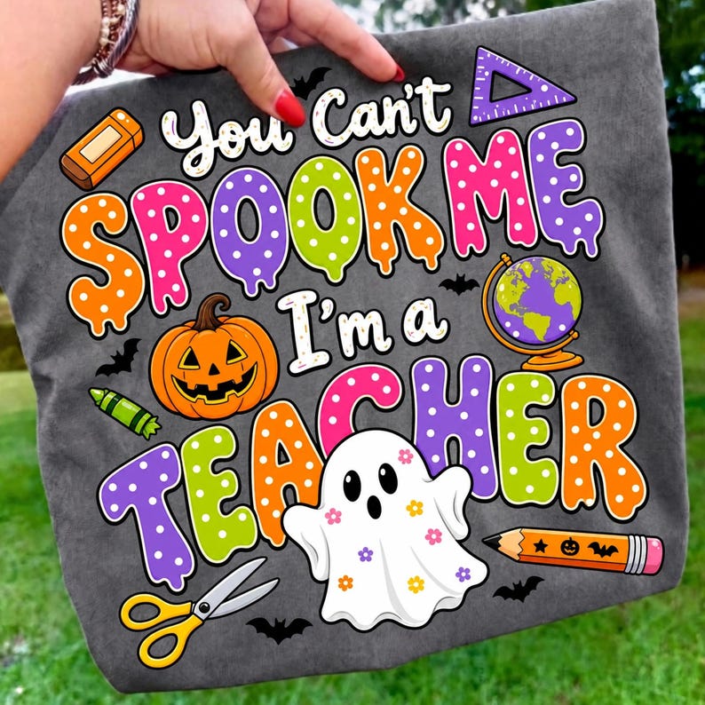 May include: A dark gray t-shirt featuring a colorful graphic with the text "You Can't SPOOK ME I'm a TEACHER". The design includes a jack-o'-lantern, a globe, a ghost with flower patterns, scissors, a pencil, an eraser, and a triangle ruler.