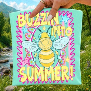 Peut inclure: Un sac fourre-tout vert menthe clair orn&eacute; d'une illustration centrale d'abeille jaune et noire entour&eacute;e d'&eacute;toiles roses et jaunes. Le texte BUZZIN INTO SUMMER est &eacute;crit en lettres jaunes grasses. Le motif inclut une bordure rose ondul&eacute;e.