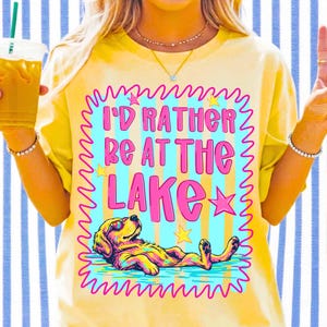 May include: A pale yellow t-shirt featuring a graphic print with the text I'D RATHER BE AT THE LAKE in pink letters. The design includes a golden retriever wearing sunglasses, floating in blue water, surrounded by pink and yellow stars.