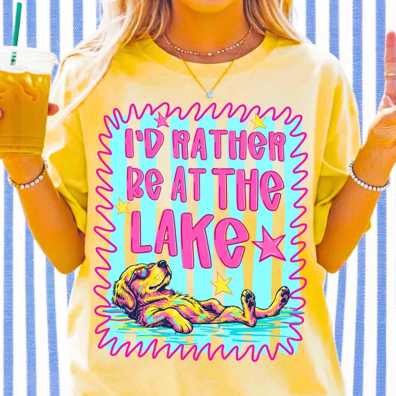 May include: A pale yellow t-shirt featuring a graphic print with the text I'D RATHER BE AT THE LAKE in pink letters. The design includes a golden retriever wearing sunglasses, floating in blue water, surrounded by pink and yellow stars.