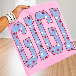 May include: A light pink fabric square featuring the word GIGI in large, block-style letters. The letters are light blue with a distressed texture and are decorated with a pattern of small red and white stars throughout the design.