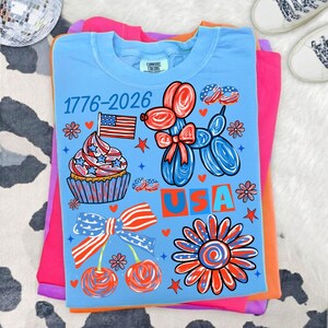 May include: A light blue t-shirt featuring a graphic print with the text 1776-2026 and USA. The design includes a cupcake with an American flag, a balloon dog, a large bow, two cherries, a flower, and small stars in red, white, and blue colours.