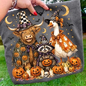 May include: A dark gray fabric featuring a graphic print of a Highland cow, a goose, and a raccoon wearing checkered witch hats. The animals are dressed in capes decorated with pumpkins and ghosts, surrounded by jack-o'-lanterns, bats, and stars.