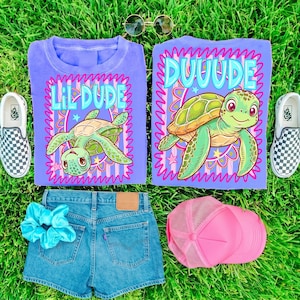 May include: Two purple t-shirts featuring a green sea turtle graphic and the text LIL DUDE and DUUUDE, blue denim shorts, a light blue scrunchie, a pink mesh trucker hat, and a pair of black and white checkered slip-on sneakers.