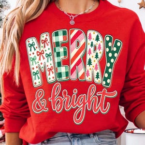 May include: A bright red crewneck sweatshirt featuring the text MERRY in large block letters, each filled with different holiday patterns including bows, green plaid, red stripes, trees, and green dots, with & bright written below in red script.