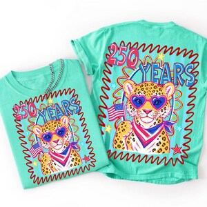 May include: A mint green t-shirt featuring a graphic of a leopard wearing heart-shaped sunglasses and a red, white, and blue bandana. The leopard holds a small American flag. The text 250 YEARS is printed above the leopard in blue and red.