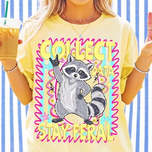 Op de afbeelding: Een lichtgeel t-shirt met een afbeelding van een grijze wasbeer die een rock-and-roll handgebaar maakt. De tekst luidt COLLECT DATA en STAY FERAL in gele letters, omringd door een felroze golvende rand en lichtblauwe verticale strepen.
