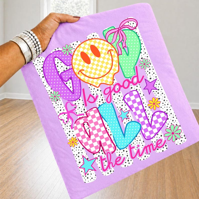 May include: A light purple fabric featuring a graphic print with the text God is good all the time. The design includes a yellow checkered smiley face, pink and blue checkered letters, black polka dots, green shapes, and small flowers.