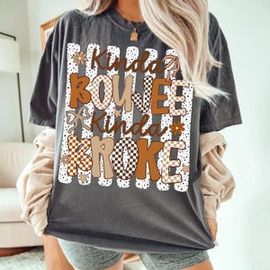 Pode incluir: Uma t-shirt cinza-carv&atilde;o de manga curta com uma estampa gr&aacute;fica de listras verticais brancas e pontos pretos. O texto Kinda BOUJEE Kinda BROKE apresenta padr&otilde;es xadrez e s&oacute;lidos em tons de castanho e bege, com flores, estrelas e la&ccedil;os.