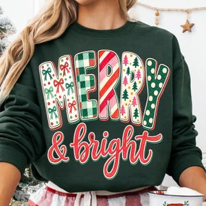 May include: A dark green long-sleeve sweatshirt featuring the text MERRY in large block letters with varied holiday patterns including red bows, green plaid, red and white stripes, pine trees, and green polka dots, with & bright in red script.