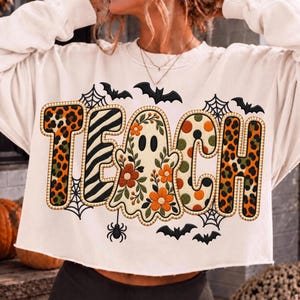 May include: A cream-colored long-sleeve sweatshirt featuring the word TEACH in large, patterned letters. The T has an orange leopard print, the E has black and white zebra stripes, the A is a ghost with orange flowers, the C has orange dots, and the H has leopard print. Black bats and spiderwebs surround the text.
