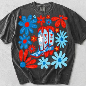 K&ouml;nnte beinhalten: Ein anthrazitfarbenes Kurzarm-T-Shirt mit einer zentralen Grafik aus zwei Cowboystiefeln, die mit einem wei&szlig;en Stern und roten Herzen verziert sind. Die Stiefel sind von stilisierten blauen und roten Blumen auf blauem Grund umgeben.