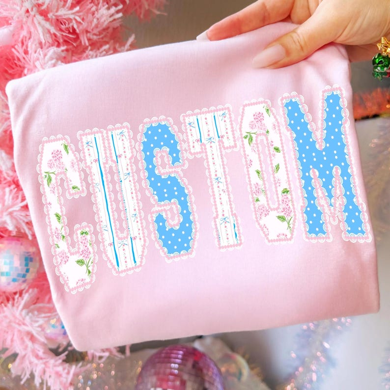 May include: A light pink t-shirt with the word "CUSTOM" in a decorative font. The letters have a white lace trim and are filled with floral and polka dot patterns. The shirt is held by a hand.