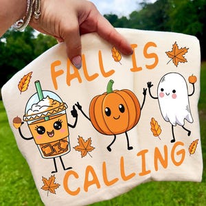 Pu&ograve; includere: Una t-shirt color crema con una stampa grafica che riporta il testo FALL IS CALLING. Il design include un caff&egrave; freddo, una zucca sorridente e un fantasma felice, circondati da piccole foglie autunnali arancioni e una piccola ghianda.