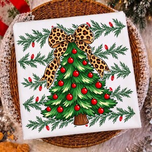 Op de afbeelding: Een wit t-shirt met een afbeelding van een groene kerstboom versierd met rode kerstballen en bekroond met een grote strik met luipaardprint. Het achtergrondpatroon bestaat uit groene dennentakken en kleine rode lichtjes verspreid over het shirt.