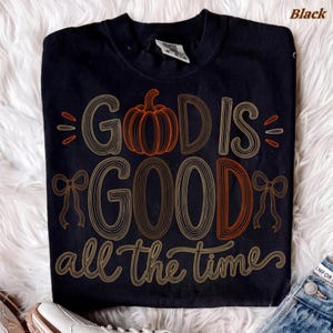 May include: A black t-shirt featuring the text "GOD IS GOOD all the time" in a stylized, multi-line font. The letter "O" in "GOD" is replaced by an orange pumpkin illustration, and the design includes two decorative line-art bows on the sides.