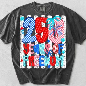 May include: A charcoal gray t-shirt featuring a graphic with the text 250 YEARS OF FREEDOM. The design includes red, white, and blue checkered patterns, firework illustrations, and light blue bows against a background of vertical white stripes.
