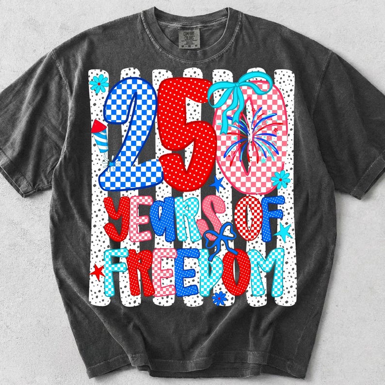 May include: A charcoal gray t-shirt featuring a graphic with the text 250 YEARS OF FREEDOM. The design includes red, white, and blue checkered patterns, firework illustrations, and light blue bows against a background of vertical white stripes.