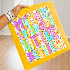 May include: A mustard yellow blanket featuring a graphic print with the text Mama's Expensive Little Bestie. The design includes colorful checkered letters in purple, blue, green, and yellow, accented with small pink bows, flowers, and a smiley face.