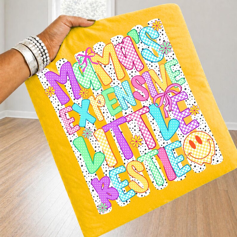 May include: A mustard yellow blanket featuring a graphic print with the text Mama's Expensive Little Bestie. The design includes colorful checkered letters in purple, blue, green, and yellow, accented with small pink bows, flowers, and a smiley face.