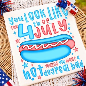 May include: A white t-shirt featuring a graphic of a blue hot dog bun with a red hot dog and white wavy line. The text reads: YOU LOOK LIKE THE 4TH OF JULY MAKES ME WANT A HOT DOG REAL BAD. Red and blue stars surround the central graphic design.