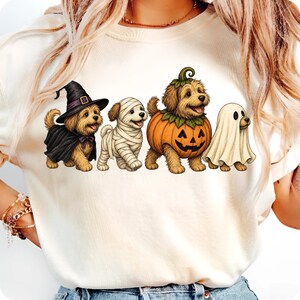 Puede incluir: Una camiseta de color crema con un gr&aacute;fico de cuatro perros peque&ntilde;os con disfraces de Halloween. Los perros est&aacute;n vestidos de bruja con sombrero y capa negra, momia envuelta en vendas blancas, calabaza y fantasma con una s&aacute;bana blanca.