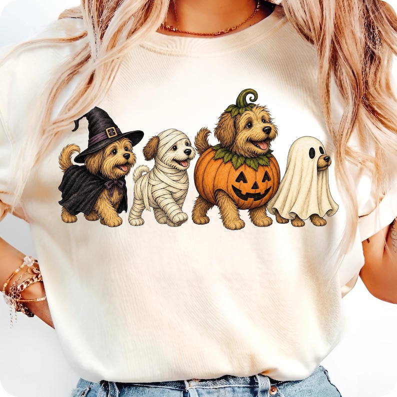 Puede incluir: Una camiseta de color crema con un gr&aacute;fico de cuatro perros peque&ntilde;os con disfraces de Halloween. Los perros est&aacute;n vestidos de bruja con sombrero y capa negra, momia envuelta en vendas blancas, calabaza y fantasma con una s&aacute;bana blanca.