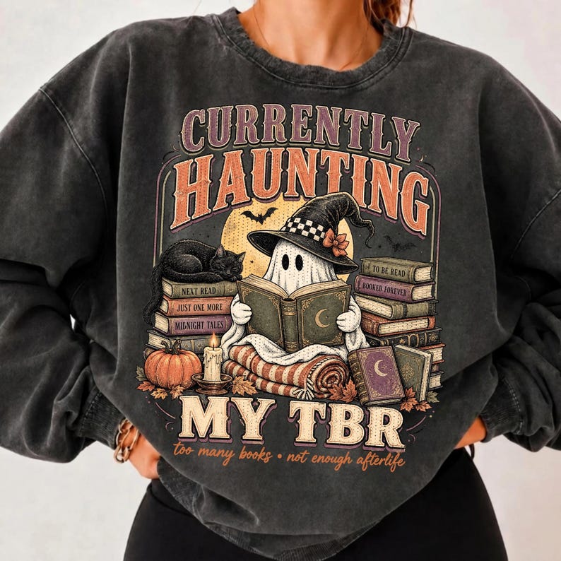 Pode incluir: Uma camisola cinzenta carv&atilde;o com lavagem mineral, apresentando um gr&aacute;fico de um fantasma com chap&eacute;u de bruxa, a ler um livro sobre uma pilha de livros. O texto diz CURRENTLY HAUNTING MY TBR e too many books not enough afterlife.