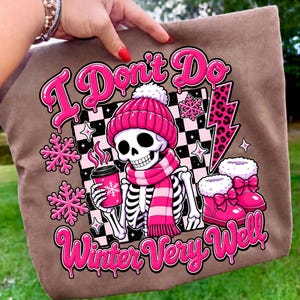 May include: A brown tote bag featuring a graphic of a skeleton wearing a pink beanie and scarf, holding a pink coffee cup. The design includes pink snowflakes, a leopard print lightning bolt, and the text I Don't Do Winter Very Well.
