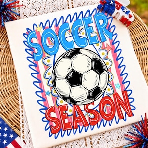 May include: A white t-shirt featuring a graphic print with the text SOCCER SEASON in blue and red. The design includes a black and white football, pink and white vertical stripes, blue wavy borders, and small yellow and blue star accents.