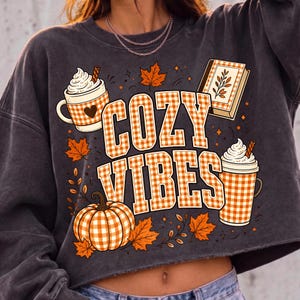 Puede incluir: Una sudadera corta de color gris carb&oacute;n con un estampado gr&aacute;fico que incluye el texto COZY VIBES en un patr&oacute;n de cuadros vichy naranja y blanco. El dise&ntilde;o presenta una calabaza, dos tazas con nata montada, un libro y hojas de oto&ntilde;o naranjas.