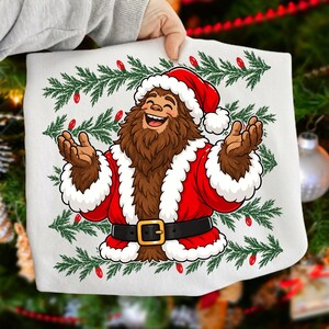 Puede incluir: Una camiseta blanca con el gr&aacute;fico de un personaje Bigfoot marr&oacute;n y peludo que viste un traje de Pap&aacute; Noel rojo con ribetes de piel blanca, un cintur&oacute;n negro con hebilla dorada y un gorro de Navidad, sobre ramas de pino verdes y luces rojas.