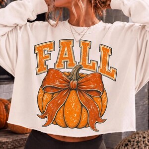 May include: A cream-colored long-sleeve sweatshirt featuring the word FALL in large orange block letters above a graphic of an orange pumpkin tied with a matching orange bow. The garment has a raw-cut hemline and a crew neckline.