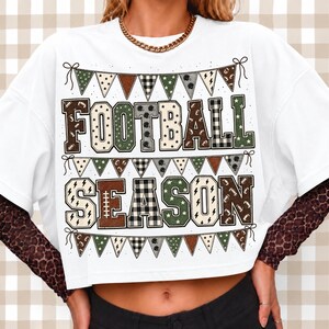 May include: A white short-sleeved t-shirt featuring a graphic print with the text FOOTBALL SEASON in block letters. The design includes rows of triangular bunting flags in brown, green, and black-and-white patterns, layered over leopard print sleeves.