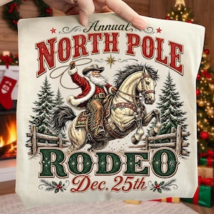 May include: A cream-colored fabric featuring a graphic of Santa Claus riding a horse while holding a lasso. The text reads Annual NORTH POLE RODEO Dec. 25th. The design includes pine trees, a wooden fence, and red and green decorative accents.