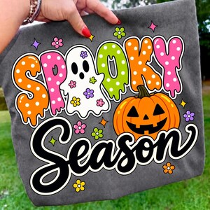 May include: A dark gray t-shirt featuring a colorful graphic with the text "SPOOKY SEASON" in a dripping, polka-dot font. The design includes a white ghost decorated with small flowers and a bright orange jack-o'-lantern pumpkin.