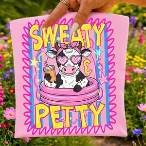 K&ouml;nnte beinhalten: Ein hellrosa Stoffquadrat mit einer Grafik einer schwarz-wei&szlig;en Kuh, die ein rosa Stirnband und herzf&ouml;rmige Sonnenbrillen tr&auml;gt und mit einem Getr&auml;nk in einem rosa aufblasbaren Pool sitzt. Der Text lautet SWEATY & PETTY in gelben Buchstaben.