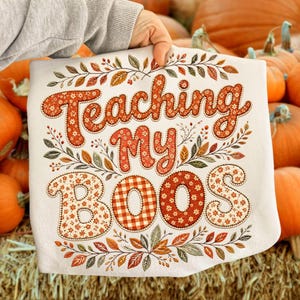 Pu&ograve; includere: Una t-shirt bianca con la scritta Teaching My BOOS in lettere arancioni e marroni. Le lettere sono riempite con motivi floreali e a quadretti, circondate da un bordo decorativo di foglie autunnali e piccole bacche dai colori autunnali.