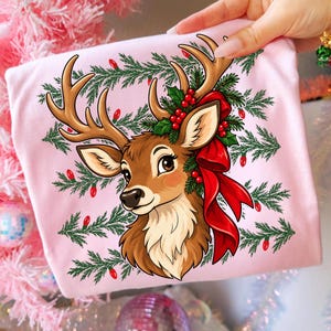 May include: A light pink t-shirt featuring a graphic illustration of a brown deer with large antlers. The deer is adorned with a red ribbon, green pine branches, and red berries. The background includes a pattern of green pine branches and red lights.