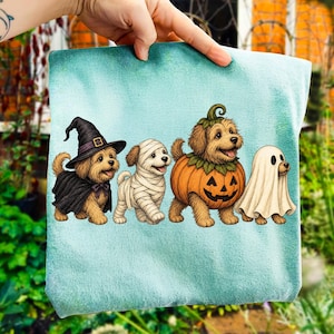 Puede incluir: Una camiseta azul claro con un gr&aacute;fico de cuatro perros peque&ntilde;os con disfraces de Halloween. Los perros est&aacute;n vestidos de bruja con sombrero y capa negra, momia envuelta en vendas blancas, calabaza y fantasma cubierto con una s&aacute;bana blanca.