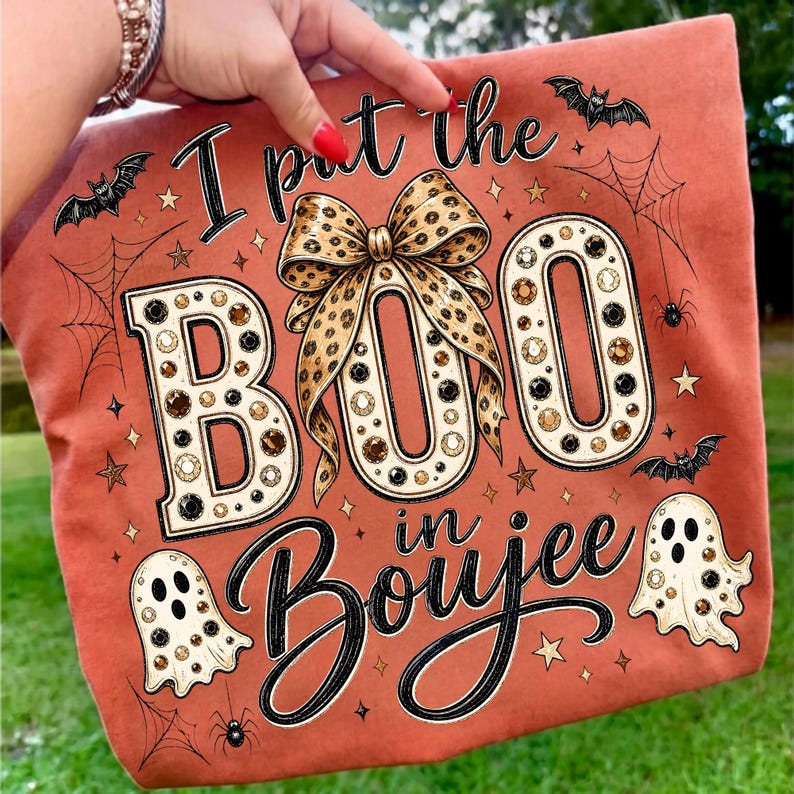 Pu&ograve; includere: Una t-shirt color arancio bruciato con la scritta I put the BOO in Boujee. Il design include un grande fiocco leopardato, due fantasmi, pipistrelli, ragnatele e stelle. Le lettere sono riempite con un motivo di strass marroni e neri.