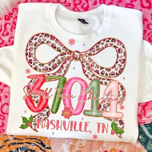 Könnte beinhalten: Weißes Sweatshirt mit festlichem Design. Es zeigt eine große Schleife mit rosa und braunem Leopardenmuster, den Text "Small Town Christmas 37014 Nashville, TN" in bunten Buchstaben und Schneeflocken-Akzente. Ein Bekleidungsartikel zum Thema Feiertage.