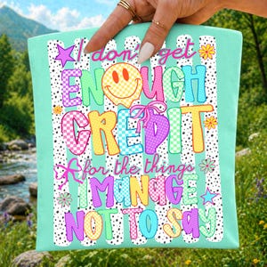 Pode incluir: Uma sacola verde-menta com uma estampa gr&aacute;fica com o texto I don't get ENOUGH CREDIT for the things I MANAGE NOT TO SAY. O design inclui um rosto sorridente amarelo, la&ccedil;os cor-de-rosa, flores coloridas, estrelas e listras com pontos pretos.