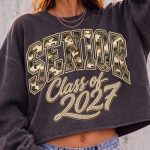 May include: A charcoal grey cropped sweatshirt featuring the word SENIOR in a green, brown, and tan camouflage print. Below the word, the text Class of 2027 is printed in a tan, script-style font with a dark outline on the front of the garment.