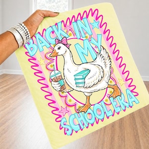 May include: A light yellow blanket featuring a cartoon goose with a pink bow, holding a drink and books. The blanket has the text "BACK IN MY SCHOOL ERA" in blue and pink. The goose is set against a pink and yellow background.