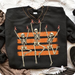 May include: A black crewneck sweatshirt featuring a graphic of three dancing skeletons. Each skeleton holds a small pink ribbon above its head. The skeletons are positioned in front of three horizontal orange brushstroke stripes.