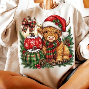 May include: A beige crewneck sweatshirt featuring a graphic of a fluffy highland cow wearing a red Santa hat and a red and green plaid scarf. The cow is surrounded by three pumpkins, pine branches, pinecones, and red holly berries.